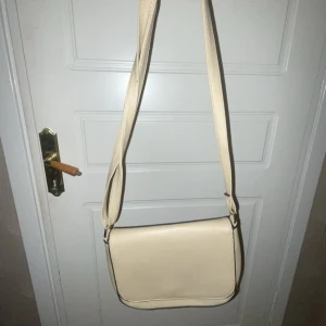 Beige axelväska från Vabene - Snygg beige axelväska från Vabene med justerbar rem och stilren design. Väskan har ett fack med dragkedja på baksidan och blommigt innerfoder med flera små fack. Perfekt storlek för mobil, plånbok och andra essentials.