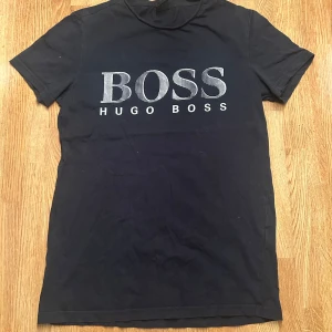Svart Hugo Boss t-shirt med logga - Svart t-shirt från Hugo Boss med stor BOSS-logga i vitt på bröstet. Klassisk rund hals och korta ärmar. Tillverkad i mjuk bomull. Perfekt för dig som gillar stilrena och enkla plagg med tydlig branding.