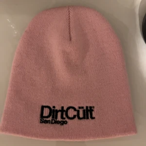 Rosa mössa från Dirt Cult - Säljer en stilren dirt cült san diego. Mössan är stickad och har en enkel, rak form med broderad svart logga framtill. Perfekt för dig som vill ha en clean look. Använd max 3gånger.