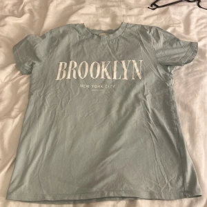 Ljusgrön t-shirt KappAhl - Fräsch ljusgrön t-shirt från KappAhl med stort vitt BROOKLYN-tryck på bröstet och texten New York City under. Klassisk rund hals och korta ärmar. Skön bomullskvalitet som passar perfekt till jeans eller shorts.