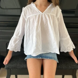 Vit blus med spetsdetaljer - Säljer en vit blus i bomull med v-ringad hals och volangkant. Blusen har vida trekvartsärmar med broderade spetsdetaljer och söt hålmönstrad kant. Perfekt för dig som gillar romantisk och chill stil. Passar till jeans eller kjol för en fräsch look. Den är något skrynklig med det går att stryka.