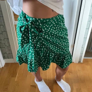 Grön blommig omlottkjol - Grön omlottkjol från ONLY med vitt blommigt mönster. Kjolen har volangkant nedtill och knyts i sidan (justerbar). Den är luftig och är i storlek S.🫶🏼