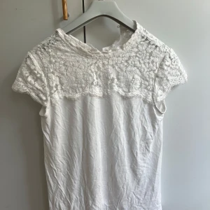 Vit spetsblus från R Édition - Supersöt vit blus från R Édition med korta ärmar och detaljerad spets upptill. Blusen har rund halsringning, knapp i nacken och blommigt spetsmönster över axlar och bröst. 