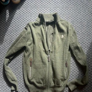 Olivgrön stickad cardigan jacka Cabbeen - Olivgrön stickad cardigan jacka från Cabbeen H.M. med dragkedja framtill och två fickor med dragkedjor. Klassisk broderad logga på bröstet och ribbade muddar. Perfekt för lager-på-lager och chill stil. Grön stickad cardigan med inga defekter och använd fåtal ggr