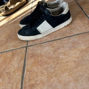 Valentino Garavani  sneakers - Snygga svarta och vita sneakers från Valentino Garavani med klassiska nitar på sulan. Skorna har svart mocka, vita skinnpaneler och svarta skosnören. Perfekt för dig som vill ha en stilren men ändå edgy look.