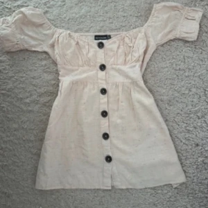 Ljus topp från PrettyLittleThing - Supersöt ljus kortklänning/topp från PrettyLittleThing med puffiga offshoulder-ärmar och stora svarta knappar framtill. har en lätt rynkad överdel och är gjord i mjuk bomull med små färgstänk. Perfekt för dig som gillar romantisk och chill stil.