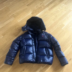 Blå dunjacka Polo Ralph Lauren XL - Snygg blå dunjacka från Polo Ralph Lauren i storlek XL. Jackan har glansig finish, huva, dragkedja och två fickor framtill. Klassisk röd Polo-logga broderad på bröstet. Perfekt för kalla dagar och riktigt skön att bära. Jackan är använd varsamt och är i nyskick. Passar xs dam. 