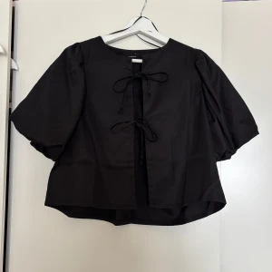 Svart blus med knyt från Gina Tricot - Supersnygg svart blus från Gina Tricot med vida, korta ärmar och två knyt framtill. Blusen har en rak, lite boxig passform och är gjord i ett mjukt bomullstyg. Perfekt för dig som gillar trendiga detaljer och enkel stil.