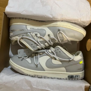 Nike Dunk Low Off-White vit/grå - Nike Dunk Low Off-White i en clean vit och ljusgrå färgway. Skorna har dubbla snörningar, Off-White-typisk text på sidan och detaljer som exposed foam på plösen. Ovandel i skinn och textil, med klassisk rund tå och platt sula. Perfekta för dig som gillar streetwear och unika sneakers.