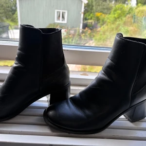 Svarta chelsea boots med klack - Stilrena svarta chelsea boots i skinnimitation med bred klack och rund tå. Resår på sidorna gör dem enkla att ta på och av. Perfekta för dig som vill ha en klassisk look med lite edge.