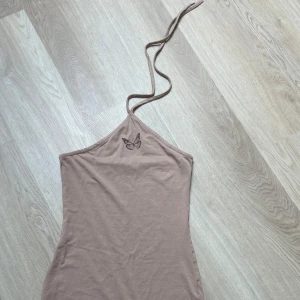 Beige kort klänning med fjäril från H&M - Säljer en beige kort klänning från H&M Divided i storlek S. Klänningen har smala axelband som knyts i nacken och en liten fjäril broderad framtill. Modellen är figurnära och ärmlös, perfekt för dig som gillar enkel och stilren design.