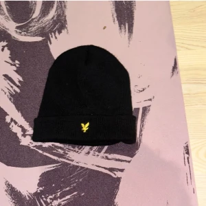 Svart mössa från Lyle & Scott - Svart ribbstickad mössa från Lyle & Scott med klassisk gul logga broderad framtill på uppviket. Enkel och stilren design som passar till det mesta. Perfekt för kalla dagar.