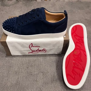 Mörkblå skor från Christian Louboutin - Snygga mörkblå skor från Christian Louboutin i mocka med ikoniska röda sulor. Skorna har rund tå, snörning och coola nitar på tån för en edgy look. Insidan är beige och sulan är vit. Perfekta för dig som vill sticka ut med exklusiv stil.
