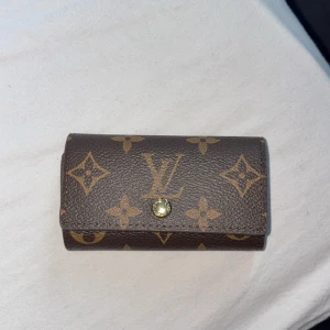 Louis Vuitton brun nyckelplånbok - Snygg brun nyckelplånbok från Louis Vuitton med klassiskt monogrammönster och guldfärgad knapp. Insidan har plats för flera nycklar.