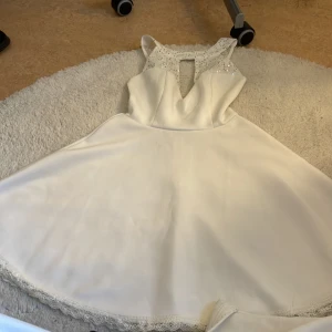 Vit kort klänning med paljetter - Supersöt vit klänning med glittrande paljetter och spets upptill. Klänningen är ärmlös och har en snygg v-ringning med öppet parti fram. Kjolen är utsvängd och har spetskant längst ner. Perfekt för student eller sommarens speciella tillfällen!