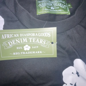 Svart blommig t-shirt från Denim Tears - Svart t-shirt från Denim Tears med stora vita blommiga tryck över hela tröjan. Klassisk rund halsringning och korta ärmar. Tillverkad i mjuk bomull som känns skön mot huden. Perfekt för dig som vill sticka ut med en unik och trendig look.