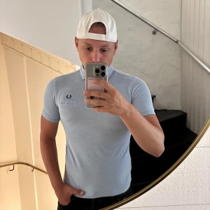 Ljusblå Fred Perry t-shirt med krage - Snygg ljusblå t-shirt från Fred Perry med klassisk krage och knappdetaljer. T-shirten har korta ärmar och en broderad Fred Perry-logga på bröstet. Materialet är mjukt och känns som bomull, perfekt för en clean och sportig look.