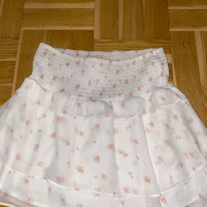 Vit blommig kortkjol från Gina Tricot - Supersöt vit kortkjol från Gina Tricot med små rosa blommor och bred smock i midjan. Kjolen har volangdetaljer.nästan inte använd  