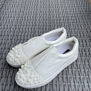 Vita sneakers med pärldetaljer Lost Ink - Unika vita sneakers från Lost Ink med coola pärldetaljer på tån. Skorna har en chunky platt sula och är gjorda i syntetmaterial med en clean look. Perfekta för dig som vill sticka ut med en trendig och lekfull stil.Vita somriga sneakers utan snörning med pärlor vid tån. Helt oanvända. Storlek 37, normal i storleken