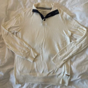 Vit pikétröja från Esprit med blå krage - Snygg vit pikétröja från Esprit med marinblå detaljer på kragen och dragkedja. Tröjan har långa ärmar, ribbade muddar och tre knappar framtill. Perfekt för dig som gillar klassisk stil med en twist. Materialet är mjuk bomull som känns skönt mot huden.