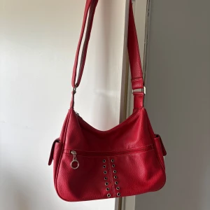 Röd axelväska med nitar och dragkedja - Cool röd axelväska i skinnimitation med bred justerbar rem. Väskan har silverfärgade nitar framtill, dragkedja med ringdetalj och en praktisk ficka på sidan.