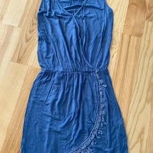 Blå omlottklänning från Rainbow XS - Supersöt blå kortklänning från Rainbow i omlottmodell med spetsdetaljer längs nederkanten. Klänningen har ärmlös design, elastisk midja och mjukt bomullsmaterial som känns skönt mot huden. Perfekt för varma dagar och chill vibes.