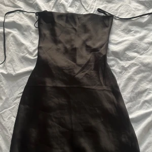 Svart satin kortklänning Miss Madison - Supersnygg svart kortklänning från Miss Madison i glansigt satinmaterial. Klänningen har tunna axelband som kan knytas och en figurnära passform som ger en elegant siluett. Perfekt för dig som vill ha en stilren och chic look med lite edge.