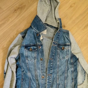 Jeansjacka med grå hoodieärmar - Snygg jeansjacka med grå sweatshirt-ärmar och huva. Klassisk blå denim framtill med bruna knappar och två bröstfickor. Perfekt för en chill streetstyle-look och passar till det mesta. Jackan har en avslappnad vibe och är riktigt bekväm.