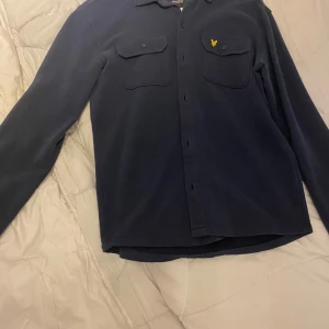 Mörkblå overshirt från Lyle & Scott - Mörkblå overshirt från Lyle & Scott med klassisk krage, två bröstfickor med lock och knappar samt den ikoniska gula loggan på vänster bröst. Skjortan har långa ärmar och stängs med knappar framtill. Perfekt för lager-på-lager-stil.