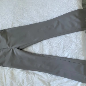 Vilma kostymbyxor grå - Snygga grå kostymbyxor med bootcut-snitt och klassisk passform. Byxorna har hällor för bälte och diskreta fickor bak. Perfekta för en clean och stilren look. Materialet känns mjukt och har en lätt glans.