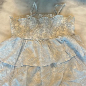 Ljusblå topp från Gina Tricot - Supersöt ljusblå topp från Gina Tricot med smock upptill och tunna axelband. Toppen har volanger och ett diskret paisleymönster som ger en chill och fräsch vibe. Perfekt för varma dagar och sommarhäng med vänner.💗bara använt 2 gånger!! Funkar som kjol också!!💞