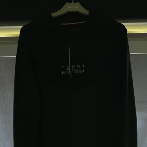 Svart hoodie Tommy Hilfiger S - Svart hoodie från Tommy Hilfiger i storlek S. Klassisk design med vit logga på bröstet, huva utan snörning och stor känguruficka framtill. Mjuk bomullskänsla och långa ärmar. Perfekt för en chill och stilren look. Ungefär 500kr nypris via booztlet, köptes 2 år sedan men som inte kom till användning mycket. Var helt enkelt inte min stil