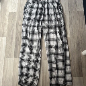 Svarta/vit rutiga pyjamasbyxor med snörning - Snygga och sköna byxor i svartvitt rutigt mönster. De har elastisk midja med snörning och raka ben. Perfekta för chill dagar eller när du vill ha en avslappnad look. Tillverkade i mjukt bomullstyg för extra komfort.