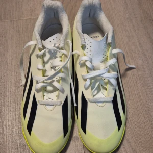 Adidas Crazyfast vita fotbollsskor - Säljer ett par Adidas Crazyfast fotbollsskor i vitt med svarta ränder och neongula detaljer. Skorna har snörning och platt sula, perfekt för inomhusspel. Lätta och smidiga med klassisk Adidas-design och texten 'Crazyfast' på sidan.