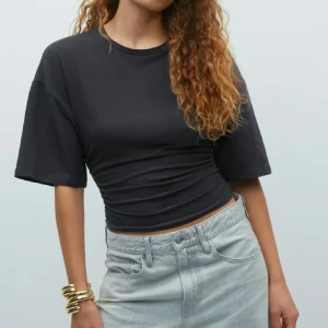 Svart croppad topp med rynk - Säljer en svart croppad topp med korta, vida ärmar och rynkade detaljer i sidan. Toppen har en rund halsringning och är i mjukt bomullsmaterial. Perfekt för en chill och trendig look.