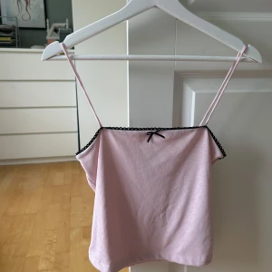 Rosa ribbad linnetopp från Gina Tricot - Supersöt ljusrosa ribbad linnetopp från Gina Tricot med tunna axelband. Toppen har svart spetskant och en liten rosett framtill som extra detalj. Perfekt för dig som gillar en söt och enkel stil. Använd någon enstaka gång. Nyskick
