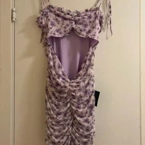 Ljuslila blommig kortklänning NLY ONE - Supersöt ljuslila kortklänning från NLY ONE med blommigt mönster i lila toner. Klänningen har smala axelband med knyt, rynkade detaljer framtill och omlott-effekt vid bysten. Materialet är mjukt och lätt polyester, perfekt för sommarens alla tillfällen. Ny kostar cirka 500 KR, helt oanvänd.