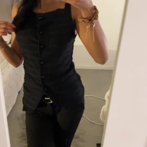 Svart linnetopp med knappar - Snygg svart linnetopp med knappar framtill och strukturerat tyg. Toppen är ärmlös och har en figurnära passform som ger en cool och stilren look. Perfekt att matcha med både jeans och kostymbyxor.