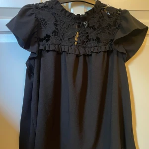 Svart spetsklänning från Emery Rose - Supersnygg svart kortklänning från Emery Rose med detaljerad spets upptill och volangkant. Klänningen har korta ärmar och en rundad hals med knäppning i nacken. Perfekt för dig som gillar romantiska detaljer och vill sticka ut med en cool look.