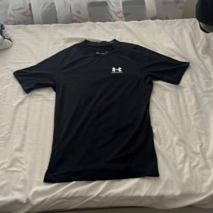 Svart kompressionst-shirt Under Armour - Svart kompressionst-shirt från Under Armour med vit logga på bröstet och ryggen. Tillverkad i ett syntetmaterial som andas, perfekt för träning. Slim fit med korta ärmar och rund hals. Snygg och sportig design som sitter tight mot kroppen.