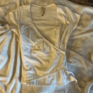 Vit omlottopp från Hunkydory - Vit omlottopp från Hunkydory med korta ärmar och v-ringning. Toppen har knyt i sidan för en snygg passform och är gjord i mjuk bomull. Perfekt för dig som gillar en clean och enkel stil med en twist.