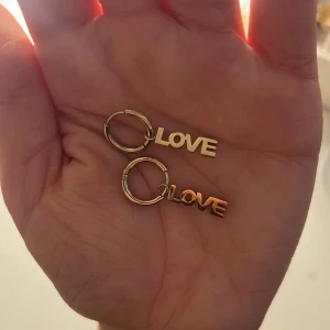 Guldiga örhängen med LOVE-hänge - Säljer ett par snygga örhängen i guldfärgad metall med texten LOVE som hänge. Runda hoops med stilren och modern vibe, perfekta för att ge din look lite extra edge och kärleksfull touch.