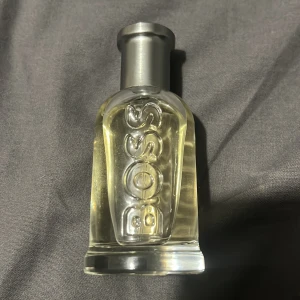 Hugo Boss Bottled parfym 100ml - Klassisk parfym från Hugo Boss i en genomskinlig glasflaska med silverfärgad metallkork där det står 'BOSS' ingraverat. Flaskan har en stilren och modern design med ljusgul vätska inuti. Perfekt för dig som gillar eleganta och fräscha dofter.
