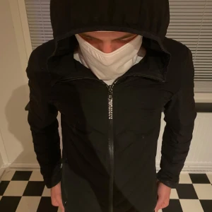 Svart pufferjacka från Emporio Armani - Svart pufferjacka från Emporio Armani EA7 med huva och dragkedja framtill. Jackan har diskret EA7-märke på ärmen och lätt quiltad design. Perfekt för dig som gillar stilren och sportig look.