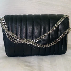 Svart quiltad axelväska med kedja - Snygg svart axelväska i quiltad design med breda vertikala sömmar. Väskan har en rejäl silverfärgad kedja som axelrem och är tillverkad i skinnimitation. Perfekt storlek för det viktigaste och ger en cool vibe till din outfit.
