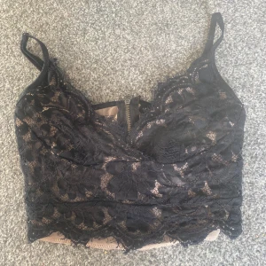 Svart spetsig croppad linnetopp - Supersnygg croppad linnetopp i svart spets med blommigt mönster och tunna axelband. Toppen har en dragkedja bak och nude-färgad underdel som ger en cool kontrast. Perfekt för att lyfta vilken outfit som helst.