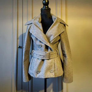 Grå kappa från ONLY  - Grå kappa från ONLY med breda slag, dubbelknäppning och ett matchande bälte i midjan. Jackan är tillverkad i ullblandning och har en klassisk, lite kortare passform med långa ärmar och markerad midja. Den är i storlek L men passar mig som vanligtvis bär M och S jätte fint.