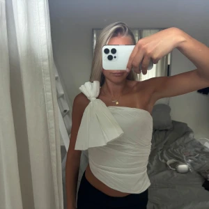 Vit oneshoulder topp med stor rosett från ZARA - Säljer en vit, asfräsch oneshoulder topp med stor rosett på axeln. Toppen är tight och croppad med draperad effekt och en asymmetrisk skärning. 🌸🌸☀️🥰