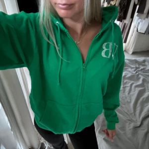 Grön hoodie från Bvaldi med strass - Grön hoodie från Bvaldi med dragkedja och huva. Stora strasstenar bildar VB-logga fram och VALDI B-text bak. Mjuk bomullskänsla, känguruficka och snörning i huvan. Perfekt för dig som gillar statement-plagg och vill sticka ut. Skulle säga storlek s/m men passar mig som xs