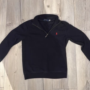 Svart half zip tröja från Polo Ralph Lauren - Svart långärmad tröja från Polo Ralph Lauren med röd broderad logga på bröstet. Tröjan har en snygg half zip-dragkedja vid halsen och är gjord i mjuk bomull. Perfekt för dig som gillar klassisk och stilren look.